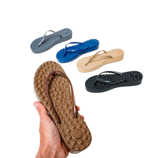 Pessoa segurando sandália Ortopédica Flyfeet Relax, mostrando a superfície com protuberâncias em alto relevo, projetada para ativar a circulação e estimular as terminações nervosas da planta dos pés, promovendo relaxamento e alívio da dor. Cores disponíveis: marrom, azul, preto, grisalho e bege. 