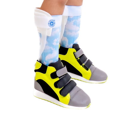 Criança usando Tênis ortopédico infantil estilo botinha, com cano alto, abertura traseira em velcro e forro acolchoado para facilitar o uso com órteses AFO ou SMO. Possui ampla abertura para encaixe do calcanhar, solado antiderrapante e estrutura que oferece suporte à postura, ajudando no tratamento de pé plano ou cavo. 
